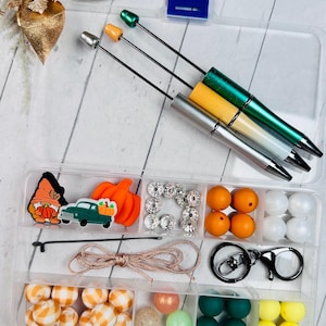 May include: A clear plastic storage box filled with various craft supplies for making fall-themed jewelry. The box includes beads in orange, white, green, and yellow, along with a gnome charm, a truck charm, a pumpkin charm, a key ring, and a spool of twine.
