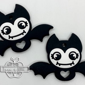 May include: Two black silicone bat charms with white faces, big eyes, and a stitched smile. Each bat has a heart-shaped cutout and a hole for hanging.