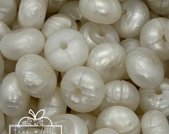 May include: Close-up of a pile of off-white, oval-shaped beads with a pearlescent sheen. Each bead has a hole running through the center. The beads are scattered, creating a textured surface. The beads are likely for jewelry making or crafts.
