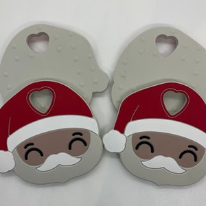 May include: Two gray silicone teethers shaped like Santa Claus. Each teether has a red hat with a white trim and a heart-shaped hole. The Santa Claus faces have black eyes, a white mustache, and a white beard.