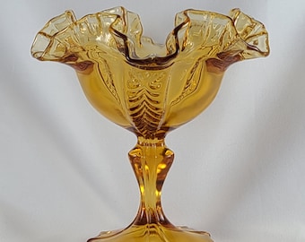 May include: Amber-colored glass pedestal bowl with a ruffled rim. The bowl has a decorative pattern on the sides and a starburst base. The stem is fluted and the bowl is likely vintage.