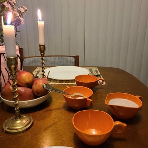 May include: A wooden table with a white plate, a bowl of pears, four orange ceramic bowls, a pitcher, and two brass candle holders with lit candles.