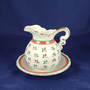 May include: A white ceramic pitcher with a green and red holly pattern and gold trim. The pitcher has a matching saucer.