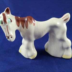 May include: A white ceramic horse figurine with brown and green painted details. The horse has a whimsical expression and is standing on its hind legs.