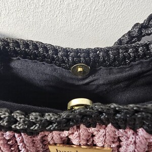May include: Close-up of a handmade crochet bag. The bag features a black top and straps, with a pink and black woven body. A small wooden tag with the word "handmade" is attached. The bag has a magnetic clasp.