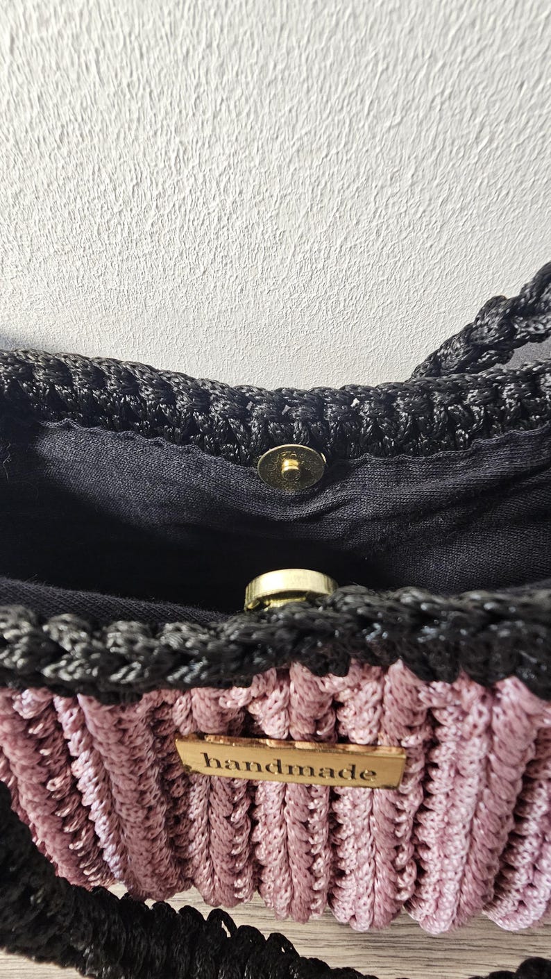 May include: Close-up of a handmade crochet bag. The bag features a black top and straps, with a pink and black woven body. A small wooden tag with the word "handmade" is attached. The bag has a magnetic clasp.