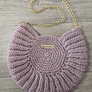 May include: A handmade, light purple crochet bag with a gold chain strap. The bag has a circular design with a textured pattern and a small rectangular tag that reads "handmade".