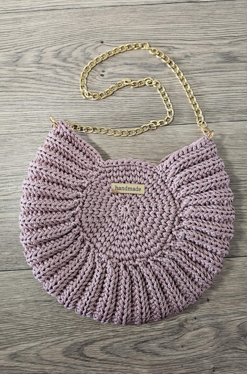 May include: A handmade, light purple crochet bag with a gold chain strap. The bag has a circular design with a textured pattern and a small rectangular tag that reads "handmade".