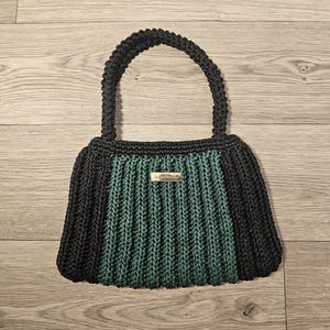 May include: A handmade black and teal crochet handbag with a sturdy handle. The bag features a rectangular shape with a teal center panel and black sides. A small metal tag that says "handmade" is attached to the front.