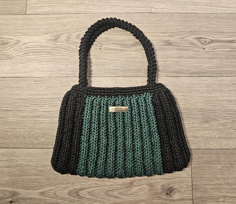 May include: A handmade black and teal crochet handbag with a sturdy handle. The bag features a rectangular shape with a teal center panel and black sides. A small metal tag that says "handmade" is attached to the front.