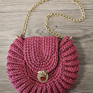 May include: A round, magenta-colored crocheted handbag with a gold-colored chain strap. The bag features a flap closure with a gold-colored clasp. The crochet pattern creates a textured, ruffled edge.