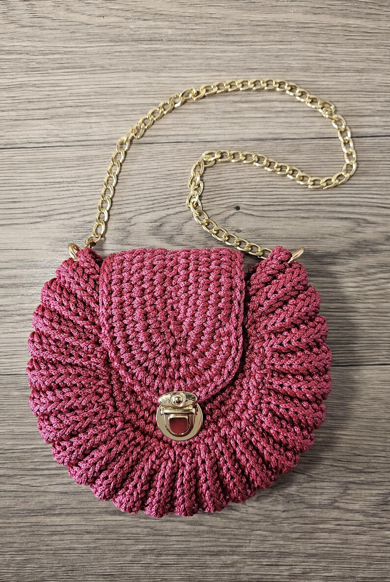 May include: A round, magenta-colored crocheted handbag with a gold-colored chain strap. The bag features a flap closure with a gold-colored clasp. The crochet pattern creates a textured, ruffled edge.