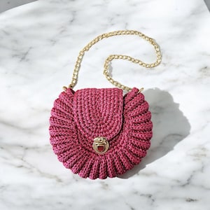 May include: A round, magenta-colored crochet handbag with a gold chain strap. The bag features a flap closure with a gold clasp. The crochet pattern creates a textured, ruffled effect around the edges, giving it a unique, handmade look.