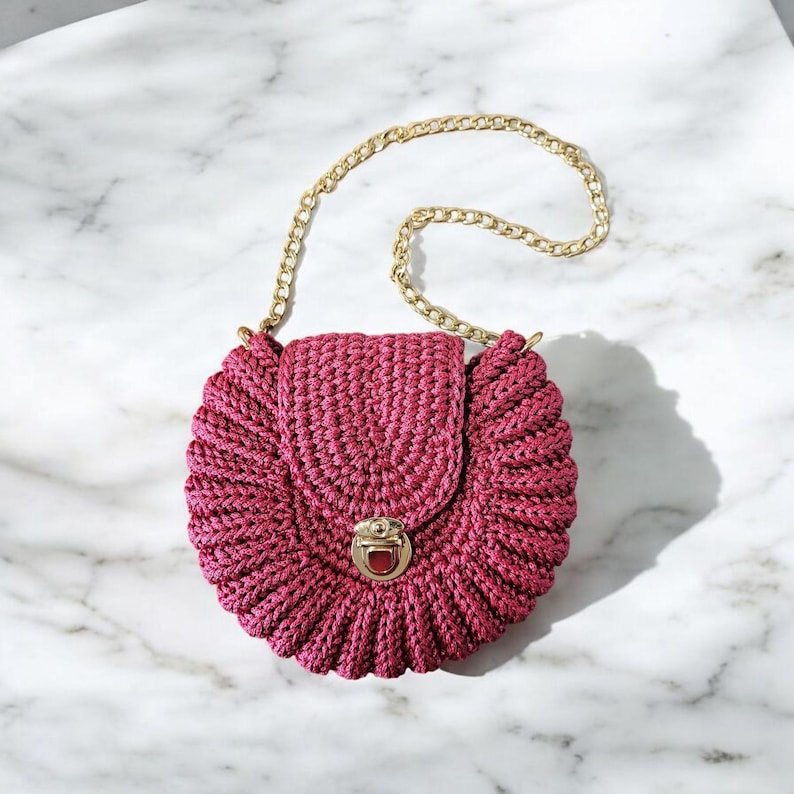 May include: A round, magenta-colored crochet handbag with a gold chain strap. The bag features a flap closure with a gold clasp. The crochet pattern creates a textured, ruffled effect around the edges, giving it a unique, handmade look.