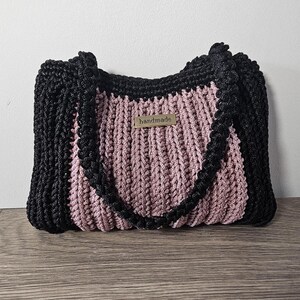 May include: A handmade tote bag with a black border and handles, and a pink center. The bag has a woven texture and a small "handmade" tag. The bag is sitting on a wooden surface.