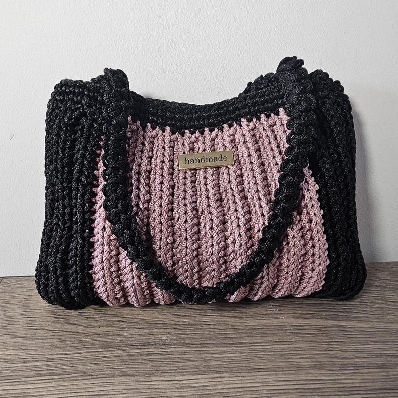 May include: A handmade tote bag with a black border and handles, and a pink center. The bag has a woven texture and a small "handmade" tag. The bag is sitting on a wooden surface.