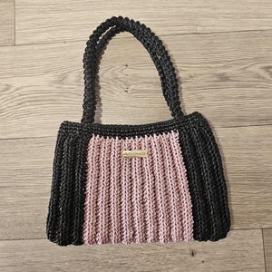 May include: A handmade black and pink crochet tote bag with two handles. The bag features a rectangular shape with a central pink panel and black sides. A small tag that says "handmade" is attached to the front.