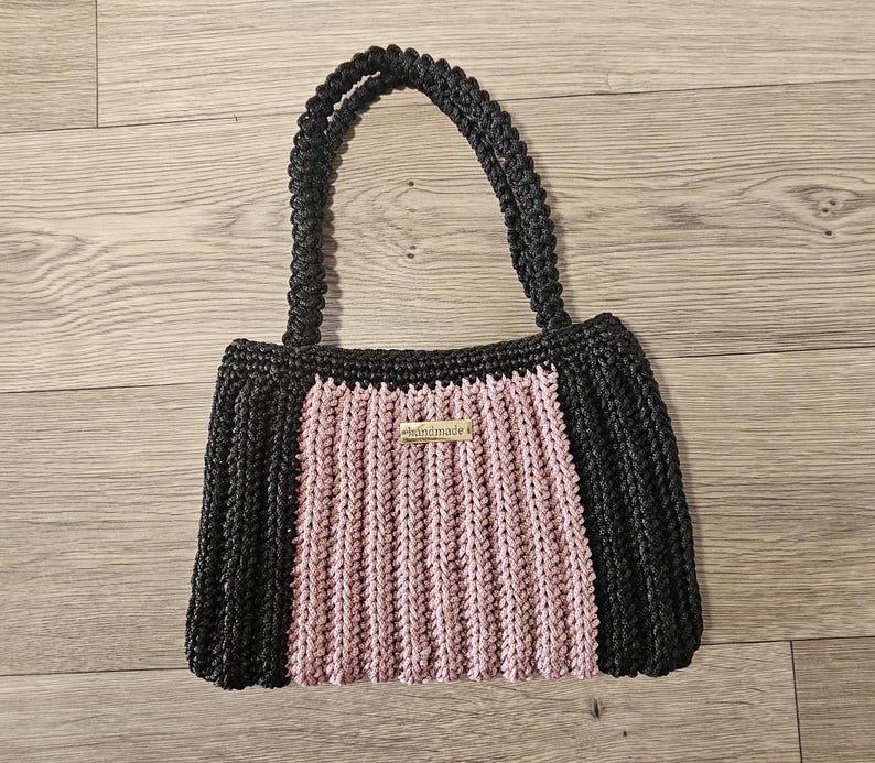 May include: A handmade black and pink crochet tote bag with two handles. The bag features a rectangular shape with a central pink panel and black sides. A small tag that says "handmade" is attached to the front.