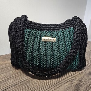 May include: A handmade black and teal green crocheted handbag. The bag features a rectangular shape with a woven pattern and a black handle. A small metal tag that reads "handmade" is attached to the front.