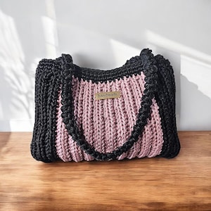 May include: A handmade tote bag with a black exterior and a pink center. The bag features a braided black handle and trim. A small, rectangular tag that reads "handmade" is attached to the front of the bag. The bag is sitting on a wooden surface.