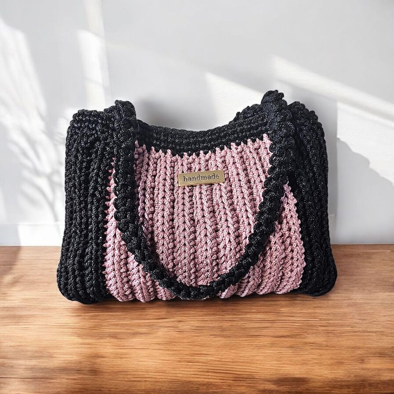 May include: A handmade tote bag with a black exterior and a pink center. The bag features a braided black handle and trim. A small, rectangular tag that reads "handmade" is attached to the front of the bag. The bag is sitting on a wooden surface.