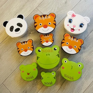 May include: A set of nine colourful lunch boxes with animal designs. The boxes are made of plastic and have a variety of animal designs, including a panda, a tiger, a frog, and a cat. The boxes are all personalised with names, including "Sophia" and "Noah".