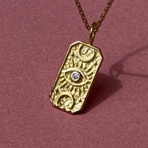 以下が含まれることがあります： 長方形のチャームが付いたゴールドカラーのペンダントネックレス。チャームは、太陽光線と三日月で囲まれた、中央に小さな透明な宝石が付いた目のデザインが特徴です。ネックレスは、ピンク色の背景に対して繊細なチェーンに掛かっています。