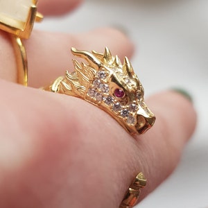 Puede incluir: Dos anillos de oro en un dedo. El anillo superior presenta una piedra blanca y el anillo inferior es una cabeza de dragón con una piedra roja para el ojo.