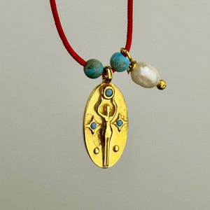May include: A gold pendant necklace with a red cord. The pendant is an oval shape with a figure design and turquoise accents. The necklace also features turquoise beads and a white pearl.