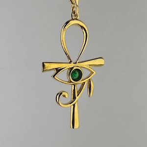 Puede incluir: Un collar con colgante dorado que presenta un símbolo ankh con un diseño de ojo. El ojo tiene una pequeña gema verde redonda en el centro. El colgante cuelga de una delicada cadena dorada, creando una joya elegante y simbólica.
