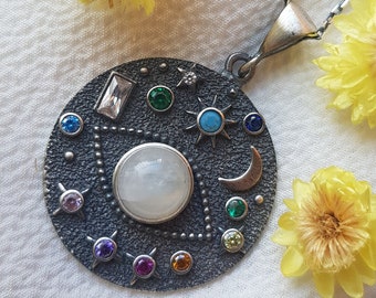 May include: A silver-toned pendant necklace with a celestial design. The pendant features a large, round, white stone in the center, surrounded by smaller colorful gemstones, a crescent moon, and sun and star motifs. Yellow flowers are scattered around the necklace.