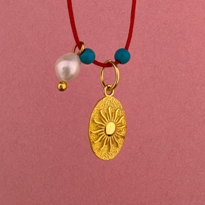 Puede incluir: Un collar con un cordón rojo fino y un colgante ovalado de tono dorado con un diseño de sol en relieve. El collar incluye una cuenta de perla blanca y dos pequeñas cuentas de color turquesa unidas al cordón cerca del colgante dorado.