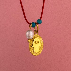 Op de afbeelding: Een ketting met een rood koord en een goudkleurige ovale hanger met een hamsa-hand symbool met een oog. De hanger is versierd met een witte parel en twee kleine turquoise kralen die aan de gouden ringetjes zijn bevestigd.