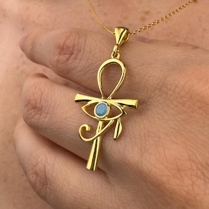 May include: A gold-colored pendant necklace featuring an Ankh symbol with an eye design. The pendant has a small, round, light blue stone in the center of the eye. The necklace is on a delicate gold chain.