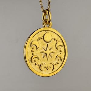 May include: A gold-toned circular pendant necklace featuring an embossed design of a central eight-pointed star surrounded by various moon phases, including crescents and a full circle, suspended from a delicate matching gold-toned chain.