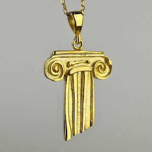 Puede incluir: Un collar con colgante dorado con un diseño de columna jónica estilizada. El colgante tiene detalles intrincados, incluyendo las volutas en la parte superior y el fuste acanalado. La cadena del collar también es dorada.