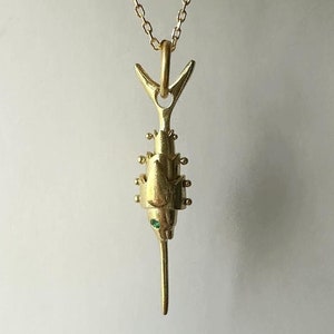 Könnte beinhalten: Eine goldfarbene Halskette mit Anhänger, die ein stilisiertes Fischdesign aufweist. Der Anhänger hat einen strukturierten Körper mit kleinen, runden Details und einem einzelnen grünen Edelsteinauge. Die Halskette ist filigran und goldfarben.