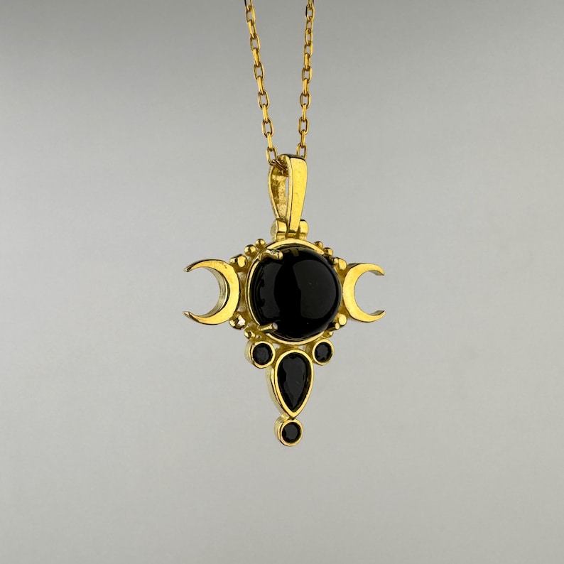 May include: A gold-colored pendant necklace featuring a central black gemstone, flanked by crescent moon shapes. Additional black gemstones are set below the central stone. The necklace hangs from a delicate gold chain.