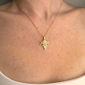 May include: A gold-colored necklace with a pendant featuring a central round, light-colored stone, flanked by crescent moon shapes. Below the central stone are two smaller, teardrop-shaped stones. The necklace is displayed on a person's chest.