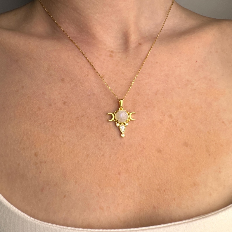 May include: A gold-colored necklace with a pendant featuring a central round, light-colored stone, flanked by crescent moon shapes. Below the central stone are two smaller, teardrop-shaped stones. The necklace is displayed on a person's chest.