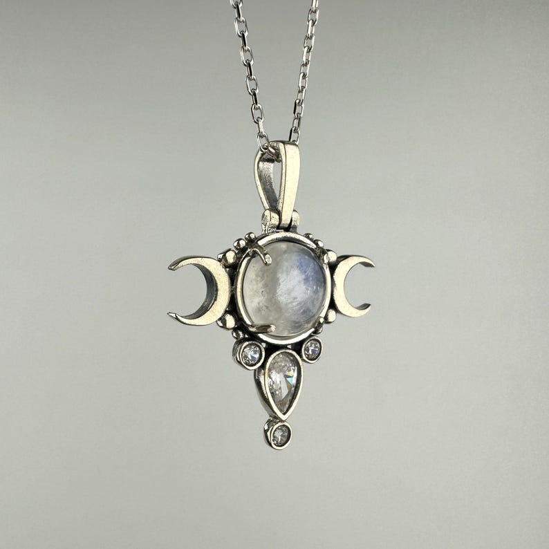 May include: A silver-toned pendant necklace featuring a central, round, iridescent moonstone, flanked by crescent moons. Below the moonstone are three clear, sparkling crystals. The pendant hangs from a delicate silver chain.
