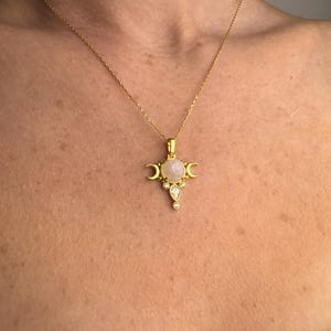 May include: Gold-toned necklace featuring a pendant with a central, round, light-colored stone, flanked by crescent moons and accented with smaller, clear stones. The necklace is displayed against a skin-toned background.