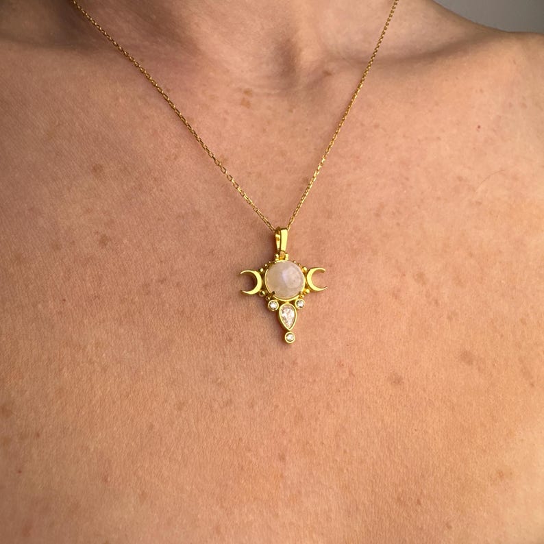 May include: Gold-toned necklace featuring a pendant with a central, round, light-colored stone, flanked by crescent moons and accented with smaller, clear stones. The necklace is displayed against a skin-toned background.