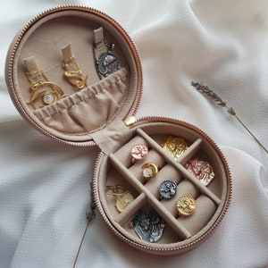 May include: A pink round jewelry box with a hinged lid. The box is open and contains a variety of gold and silver jewelry, including rings, necklaces, and earrings. The jewelry is arranged in the compartments of the box.