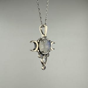 May include: A silver pendant necklace featuring a central, round, iridescent stone, flanked by three crescent moon shapes. Below the central stone are three smaller, clear, teardrop and round stones. The pendant hangs from a delicate silver chain.