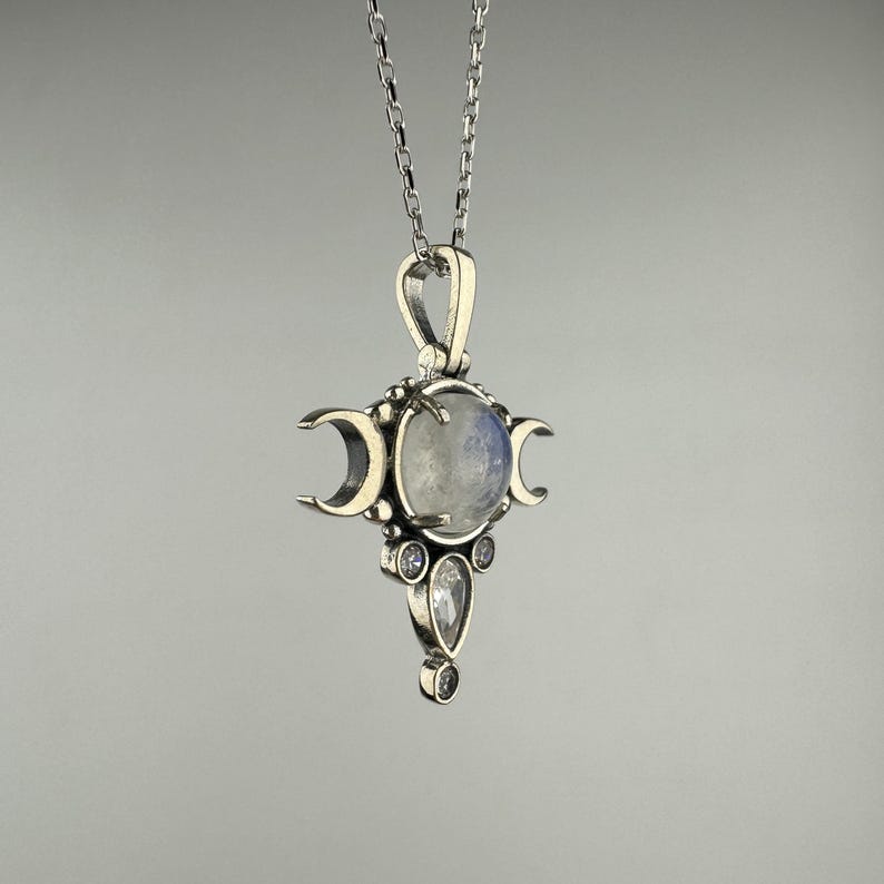 May include: A silver pendant necklace featuring a central, round, iridescent stone, flanked by three crescent moon shapes. Below the central stone are three smaller, clear, teardrop and round stones. The pendant hangs from a delicate silver chain.