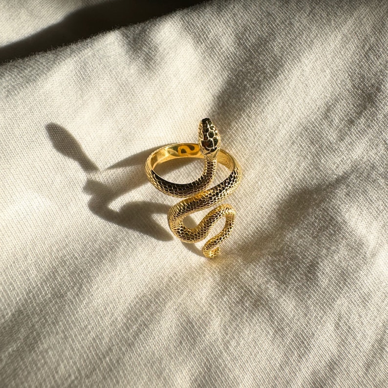 Puede incluir: Un anillo de serpiente dorado con un dise&ntilde;o en espiral. El anillo est&aacute; sobre un fondo de tela blanca.