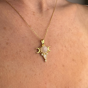 May include: Gold-toned necklace featuring a pendant with a central, round, pale gemstone, flanked by crescent moon shapes. The pendant also includes smaller, clear gemstones. The necklace is displayed against a skin-toned background.
