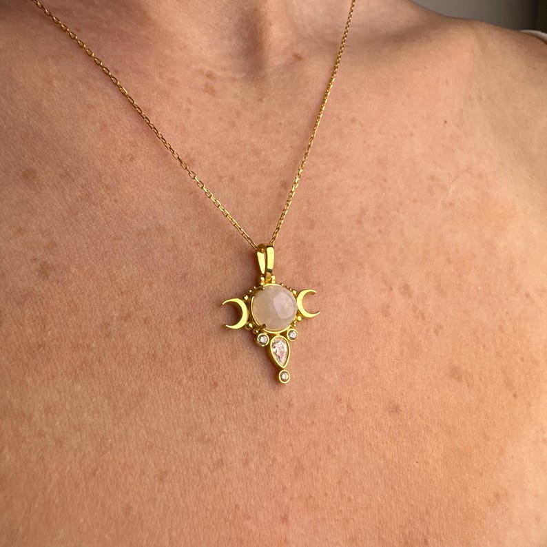 May include: Gold-toned necklace featuring a pendant with a central, round, pale gemstone, flanked by crescent moon shapes. The pendant also includes smaller, clear gemstones. The necklace is displayed against a skin-toned background.