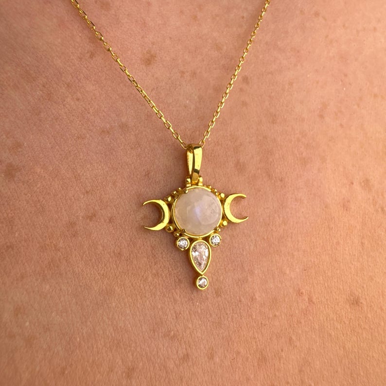 May include: A gold-colored necklace with a pendant featuring a large, round, light-colored stone at the center. The pendant has a cross-like design with crescent moon shapes and smaller, clear stones. The necklace is on a person.